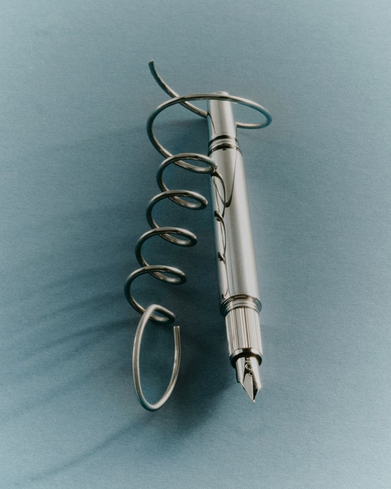 Chrome fountain pen entwined with a coiled metal wire on a muted blue-gray surface.