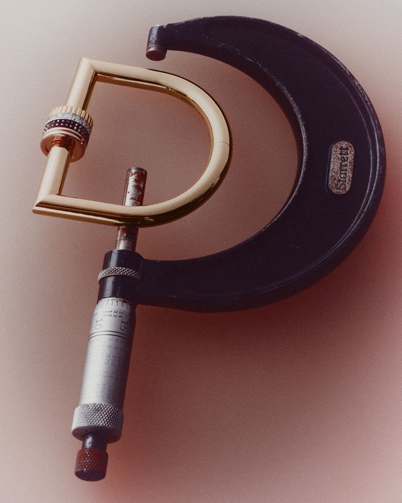 A gold D-shaped carabiner with striped gemstone bands held inside the frame of a black Starrett micrometer on a pink background.