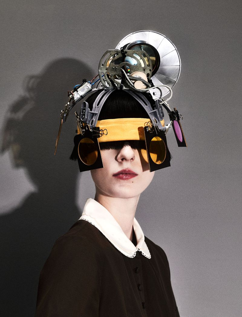 Woman wearing futuristic headgear with square lenses and silver metallic accents against a gray background. Dark dress with white collar.