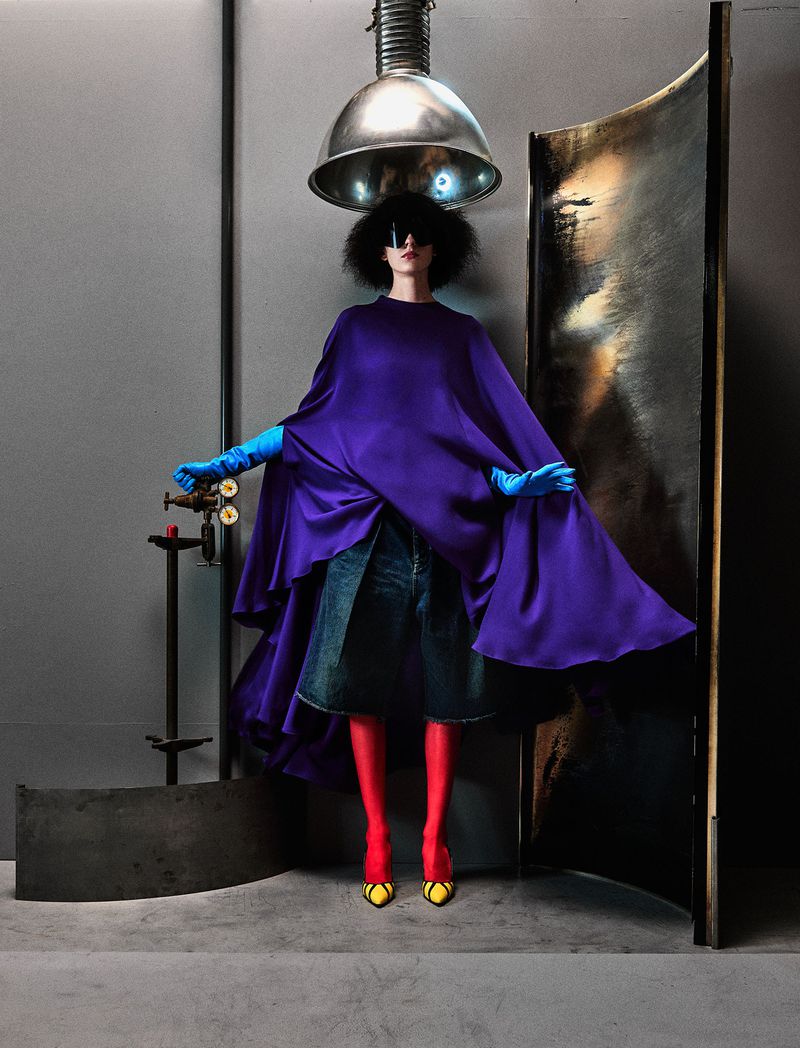 Model wearing purple cape, denim shorts, red leggings, and blue gloves beneath a large metallic dome light.