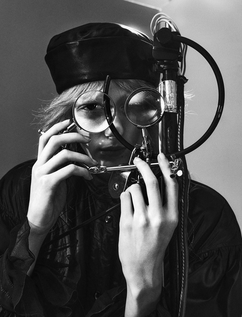 Close-up black and white portrait of a model wearing a black leather hat looking through magnifying lenses held up to their face.
