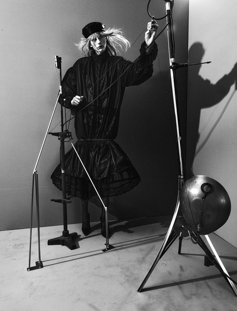 Model in a black tiered dress and cap posed among metal studio lighting stands and rigging in black and white.