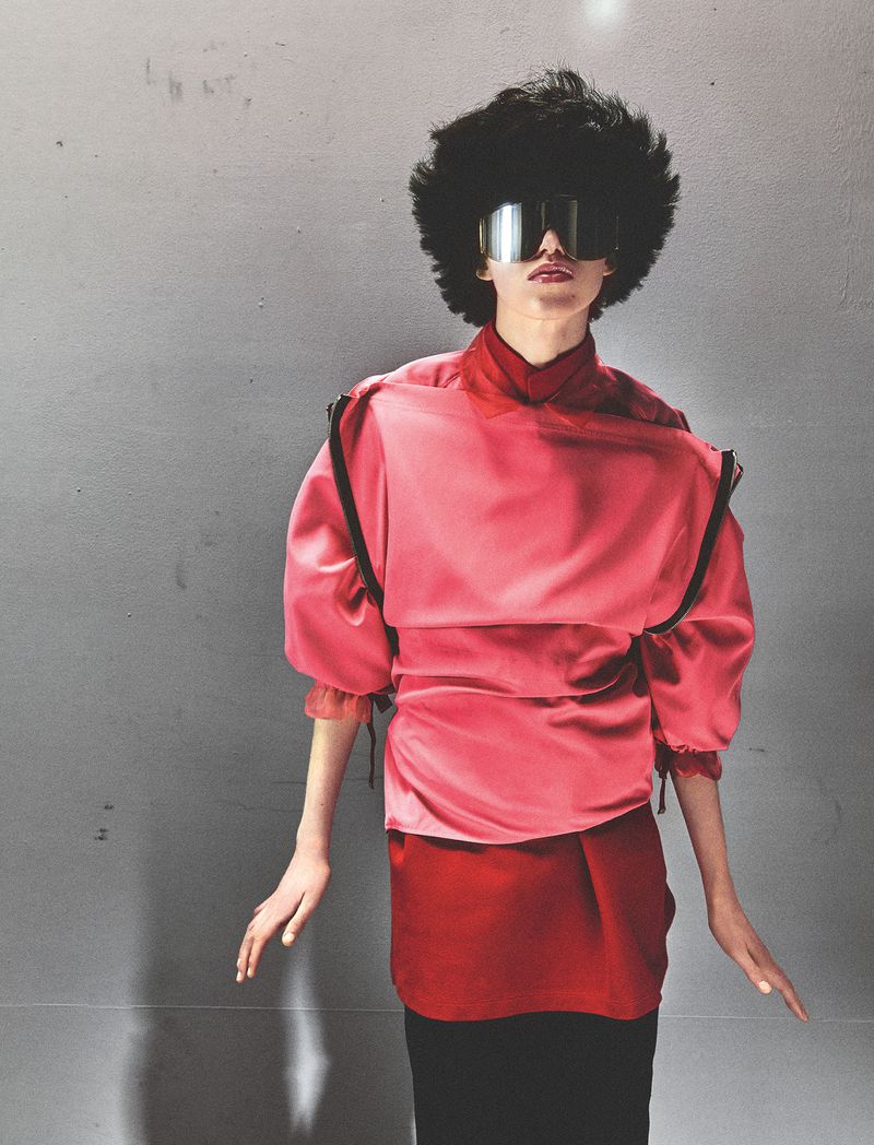 Model with voluminous dark hair wearing silver visor sunglasses, a layered red and pink tunic, and black trousers against a grey backdrop.