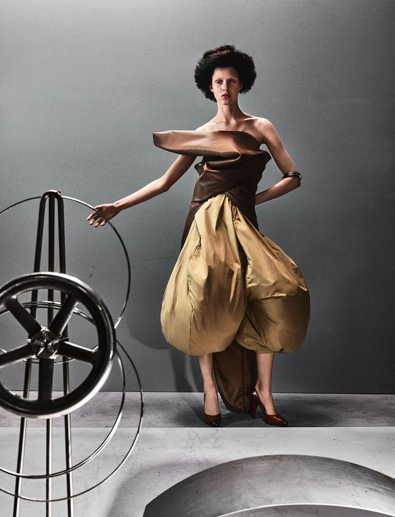 Model posing in an avant-garde brown and tan sculptural balloon dress beside a metal wheel prop against a grey studio backdrop.