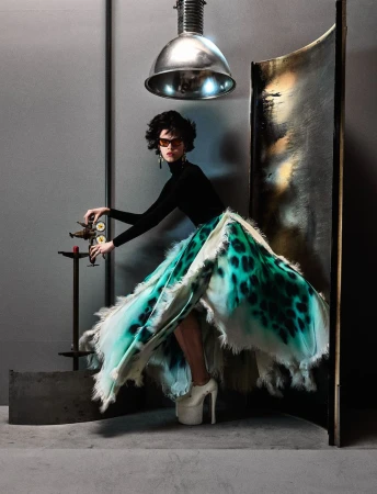 Model posing in a black turtleneck, voluminous green leopard-print ruffled skirt, and white platform heels next to industrial props.