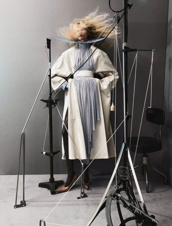 Model in an off-white coat and pleated blue dress framed by rigging and studio equipment against a grey backdrop.