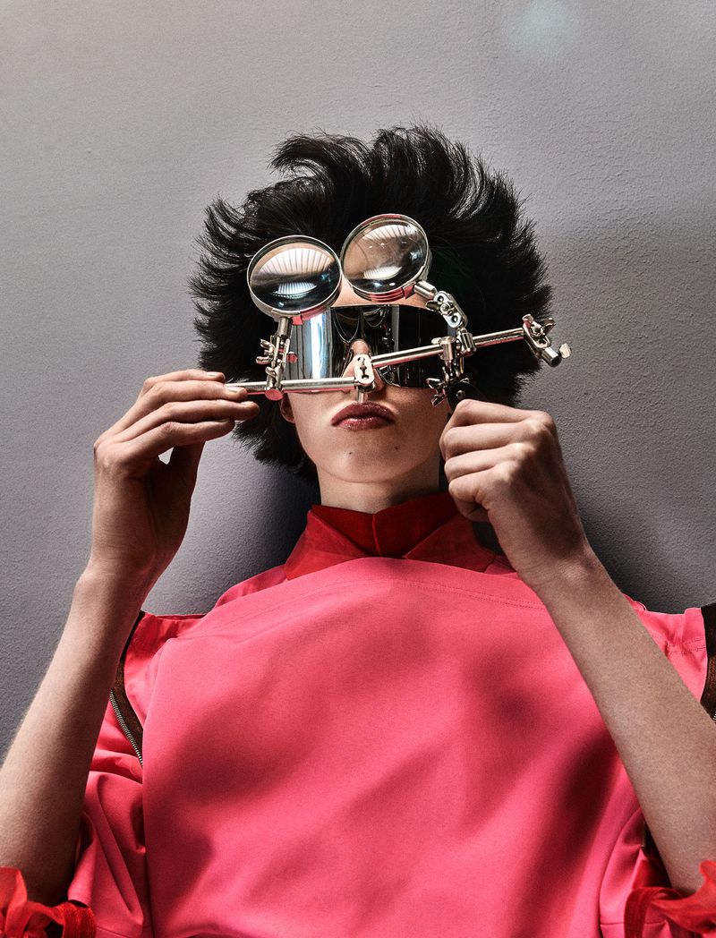 Model with dark spiky hair holding a metal magnifying glass apparatus over reflective visor glasses wearing pink against a grey backdrop.