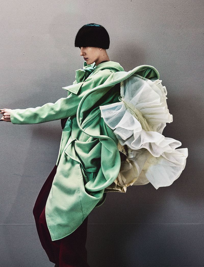 Model in profile wearing an avant-garde mint green draped coat with a voluminous white tulle back against a grey studio backdrop.