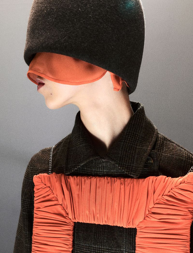 Model in profile wearing an oversized dark felt headpiece obscuring their eyes, paired with a ruched orange chest accessory.
