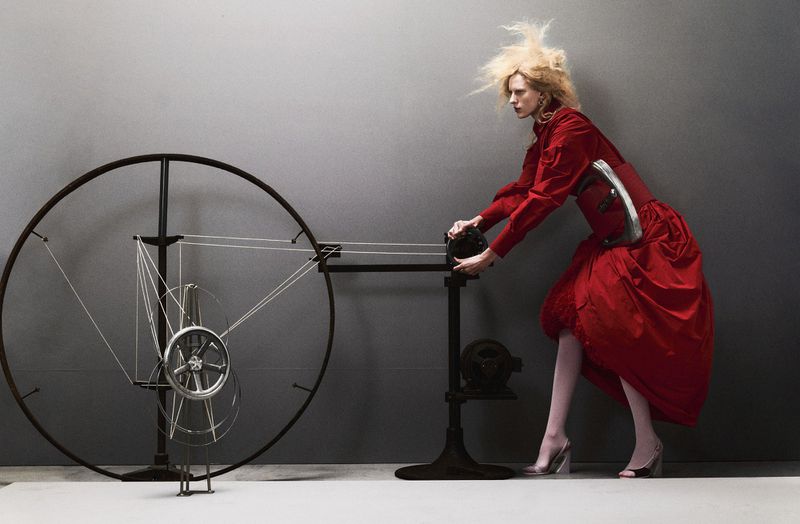 A blonde model in a voluminous red dress operates a large mechanical wire wheel apparatus against a grey studio backdrop.