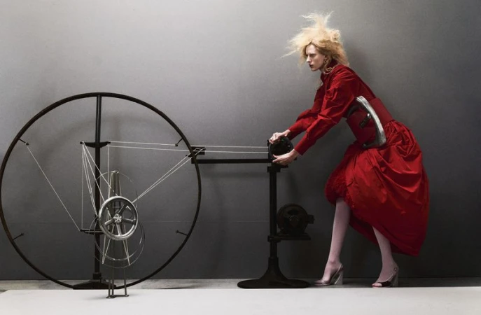 A blonde model in a voluminous red dress operates a large mechanical wire wheel apparatus against a grey studio backdrop.