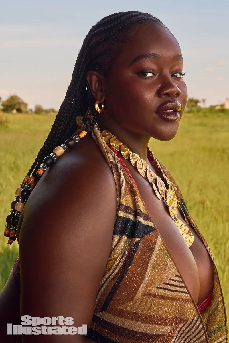 Achieng Agutu posing in a field wearing beaded braids and a gold necklace for Sports Illustrated Swimsuit.