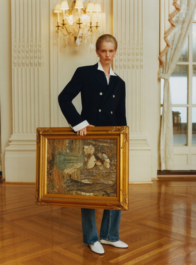 Model wearing a navy blazer and jeans holding a large ornate gold-framed painting in an elegant room with parquet floors.