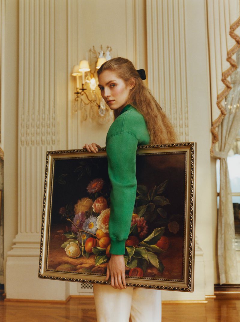 Woman in green sweatshirt holds a floral still-life painting in an elegant interior with paneled walls and chandelier lighting.