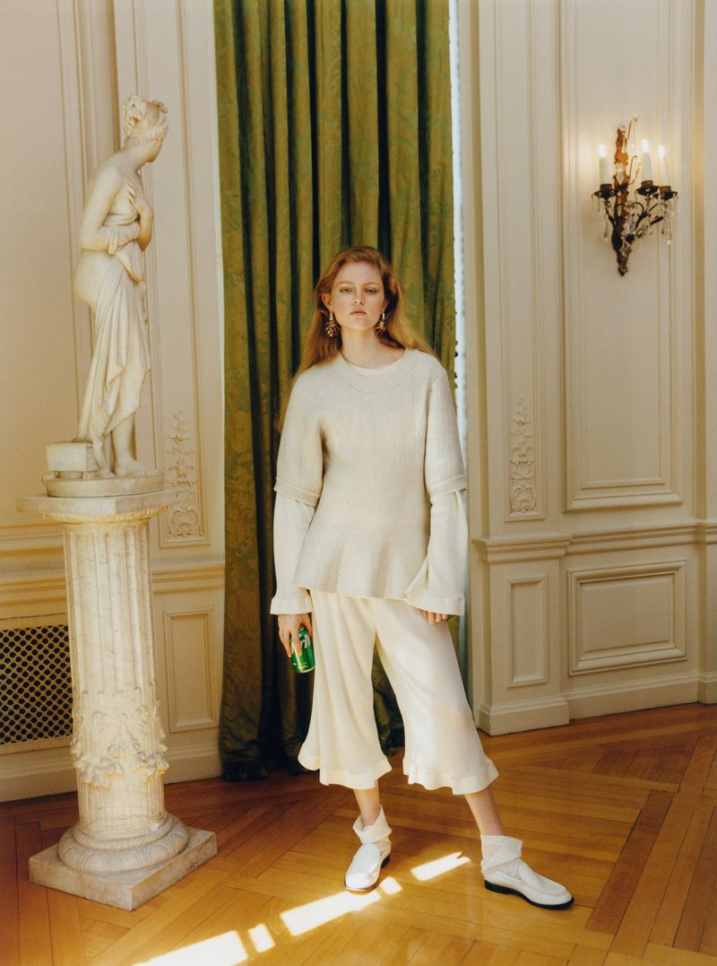 Model standing in an off-white knit sweater and culottes holding a green can next to a classical marble statue in an ornate room.