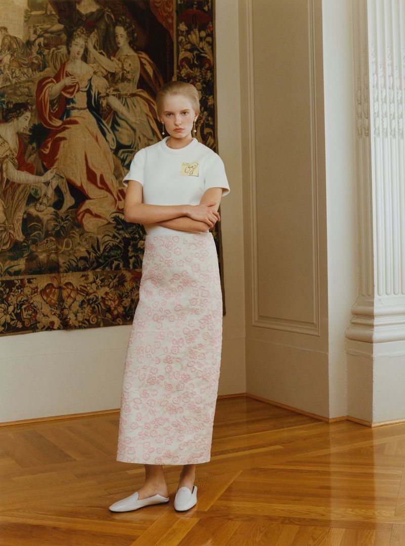 Model standing with crossed arms wearing a white short-sleeve top, textured pink column skirt, and white mules by a large tapestry.