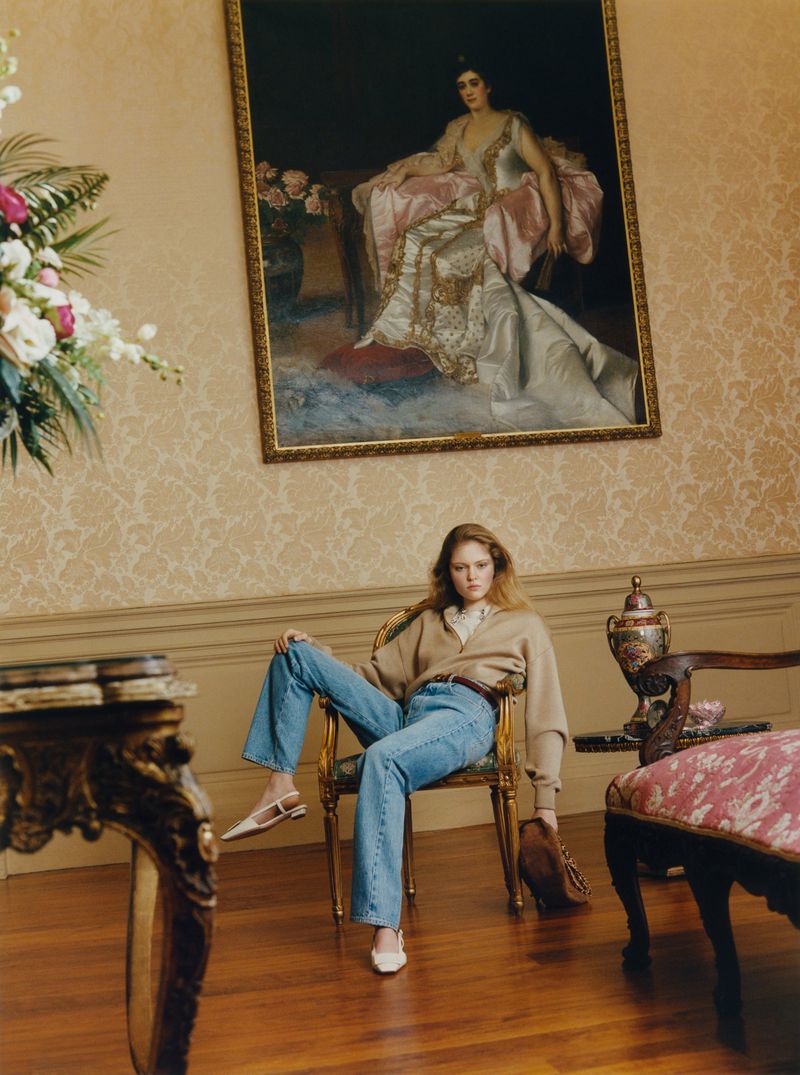 Model seated in an ornate gold armchair wearing a tan sweater, blue jeans, and white slingbacks beneath a classical portrait painting.