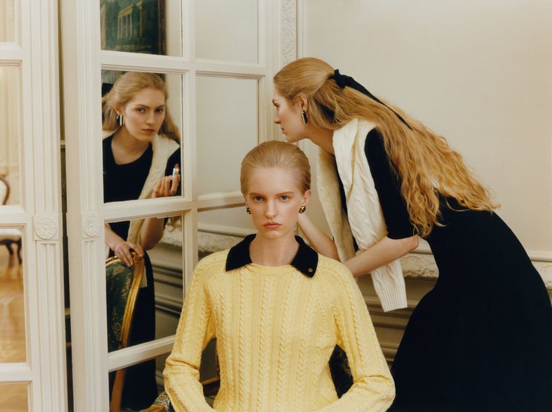 Model in a yellow cable-knit sweater seated before a paneled mirror partition with models standing behind in black and white knitwear.