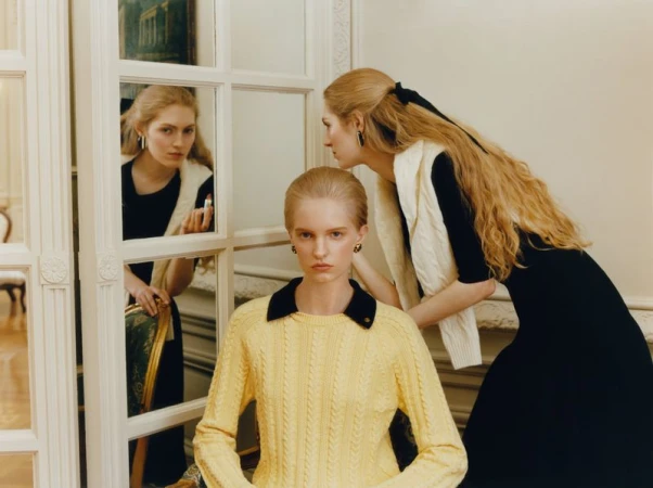 Model in a yellow cable-knit sweater seated before a paneled mirror partition with models standing behind in black and white knitwear.