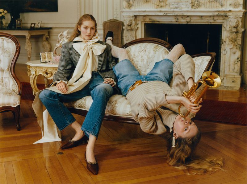 Two models pose on an antique sofa in an ornate room; one wears a blazer and giant bow while the other lies upside down holding a trumpet.