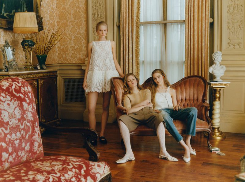 Three models posing in an ornate interior with damask wallpaper, draped curtains, and antique upholstered furniture.