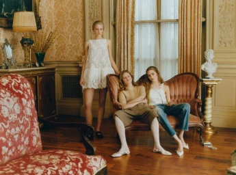 Three models posing in an ornate interior with damask wallpaper, draped curtains, and antique upholstered furniture.