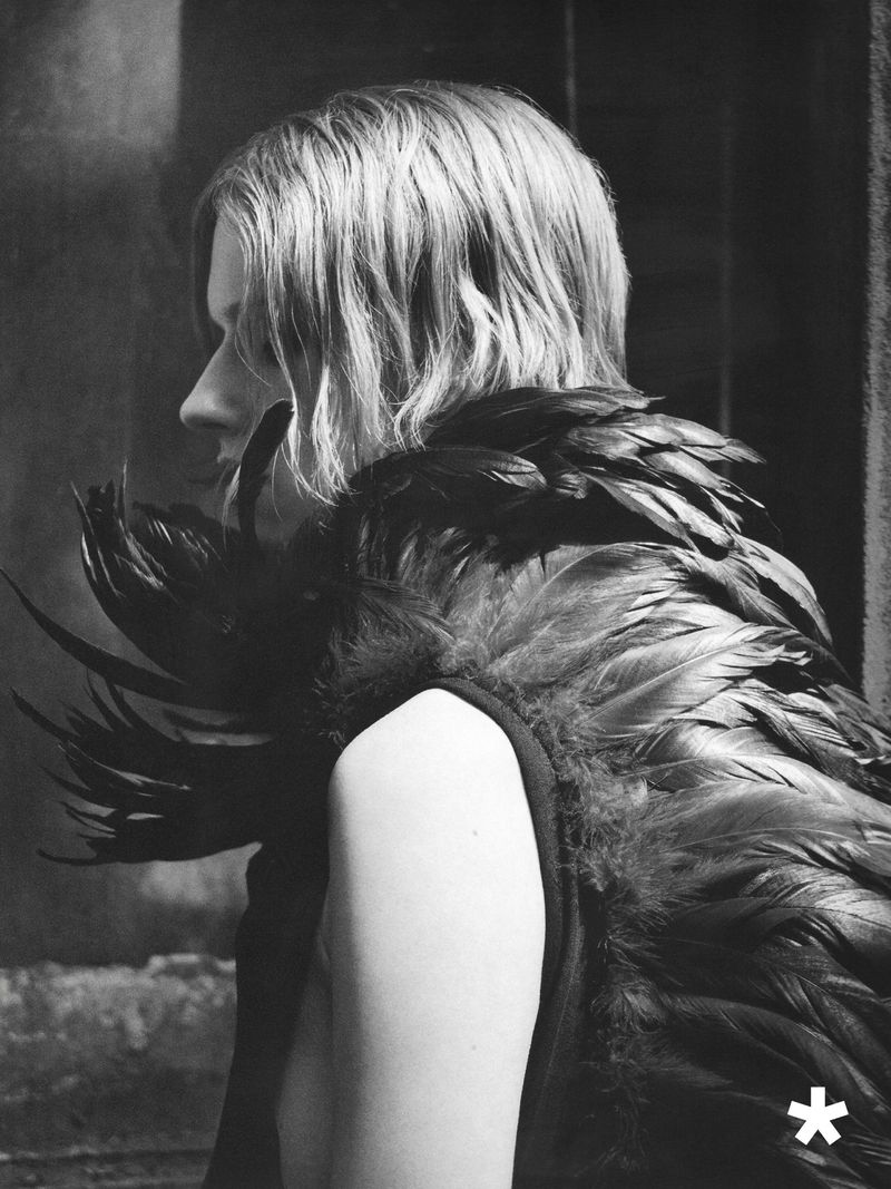 Black-and-white profile portrait of a model with wavy hair wearing an elaborate garment adorned with dark layered feathers.