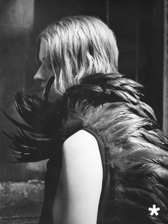 Black-and-white profile portrait of a model with wavy hair wearing an elaborate garment adorned with dark layered feathers.