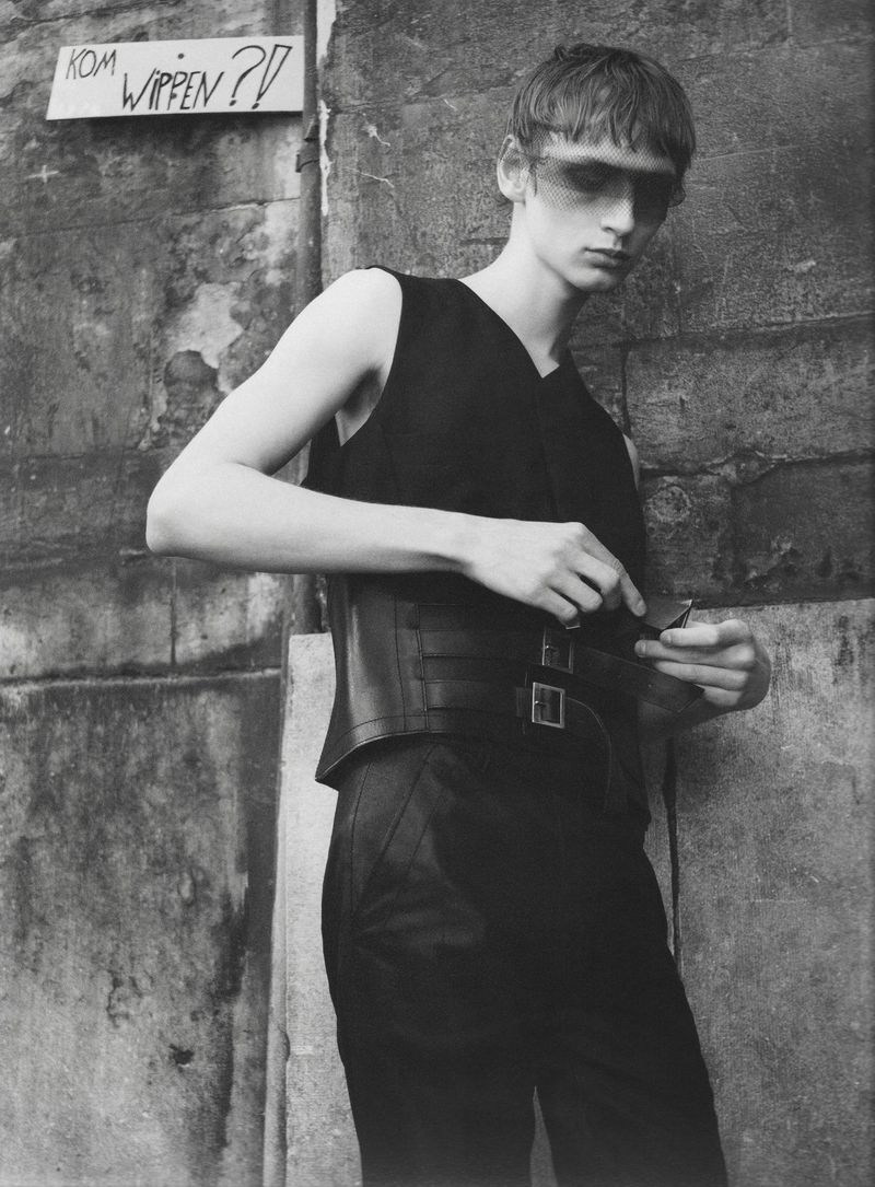 Black and white fashion portrait of a male model in a black sleeveless top and buckled corset belt standing against an aged stone wall.