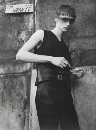 Black and white fashion portrait of a male model in a black sleeveless top and buckled corset belt standing against an aged stone wall.