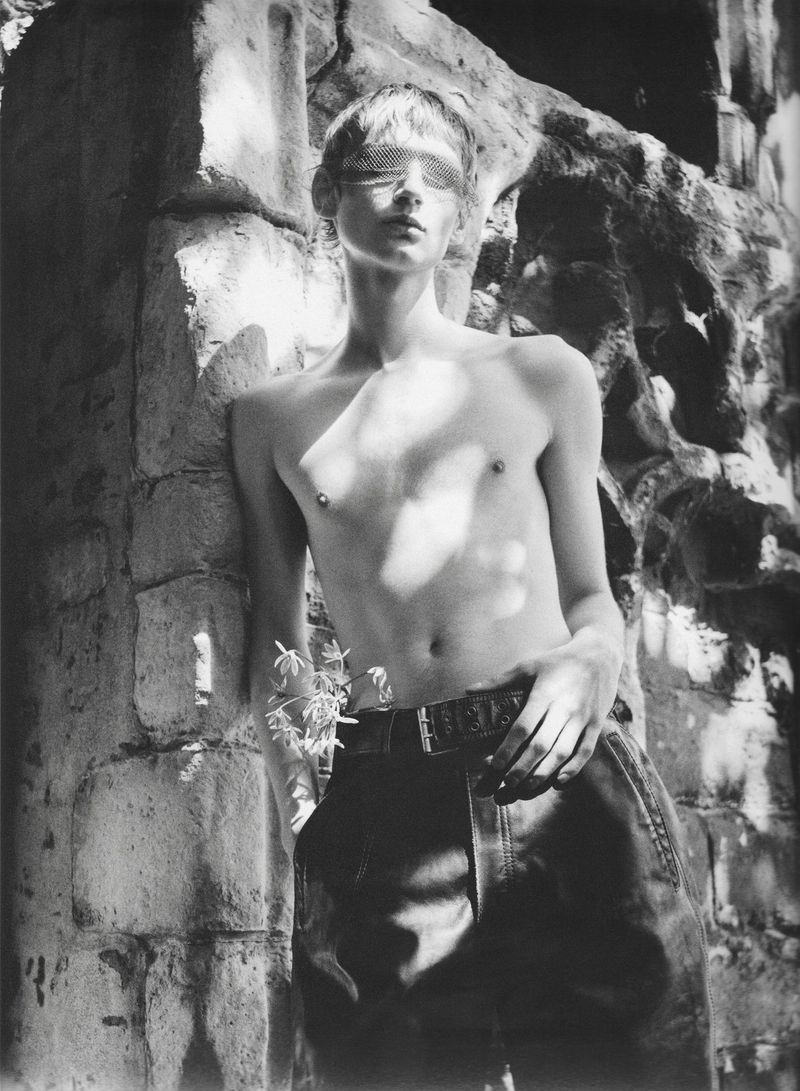 Black and white portrait of a shirtless male model wearing mesh visor eyewear and dark trousers leaning against carved stone ruins.