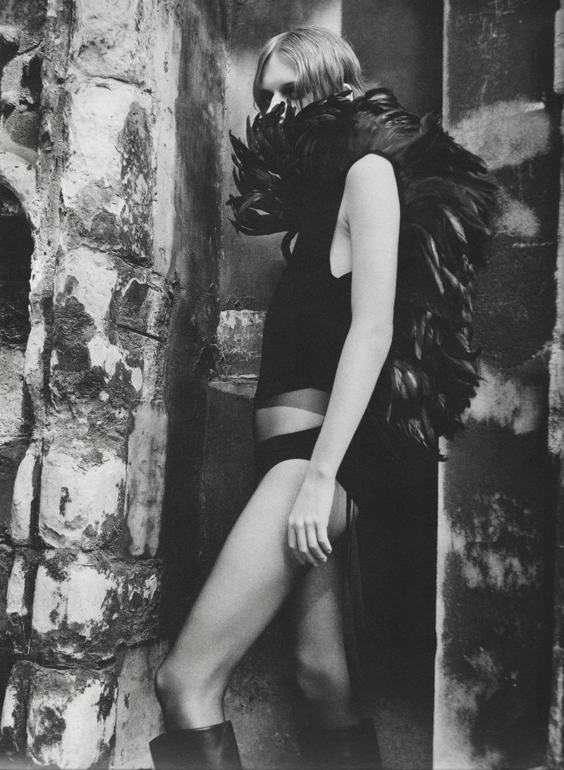 Monochrome fashion portrait of a woman wearing a voluminous black feathered collar and dark bodysuit against a distressed stone wall.