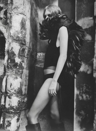Monochrome fashion portrait of a woman wearing a voluminous black feathered collar and dark bodysuit against a distressed stone wall.