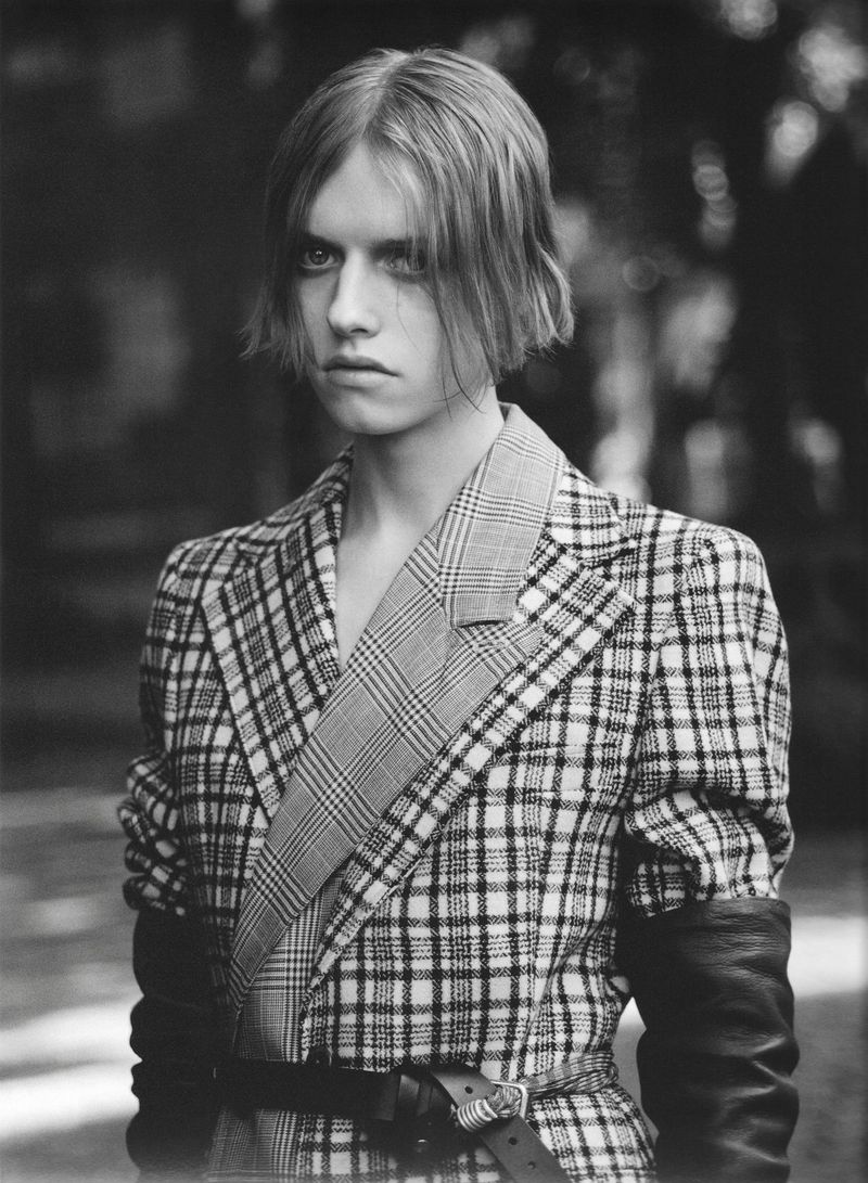 Model wearing a belted plaid tailored jacket with black leather gloves outdoors in a black and white portrait.