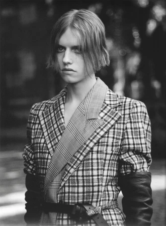 Model wearing a belted plaid tailored jacket with black leather gloves outdoors in a black and white portrait.