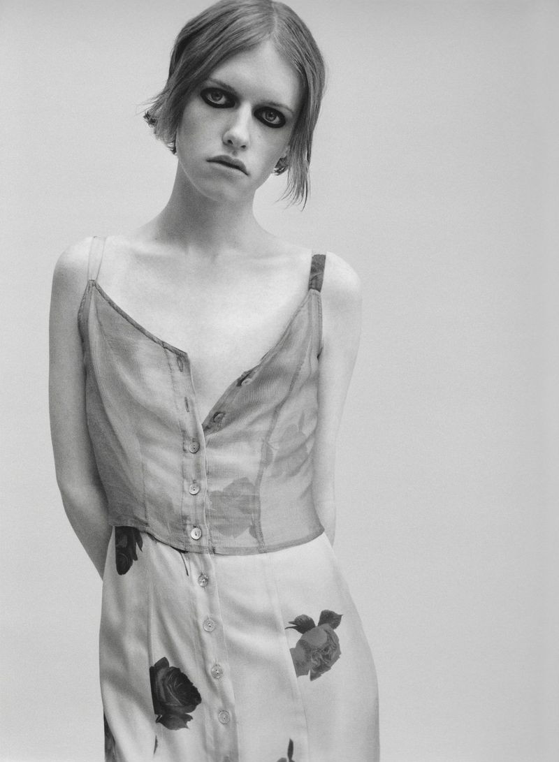 Model with dark smokey eye makeup wearing a sleeveless rose-printed button-front dress against a pale seamless background.