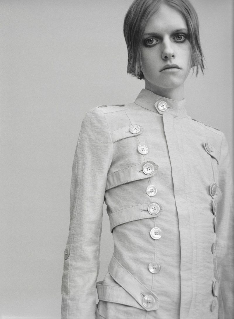 Black and white portrait of a model wearing a high-collar structured jacket with horizontal button straps against a seamless backdrop.