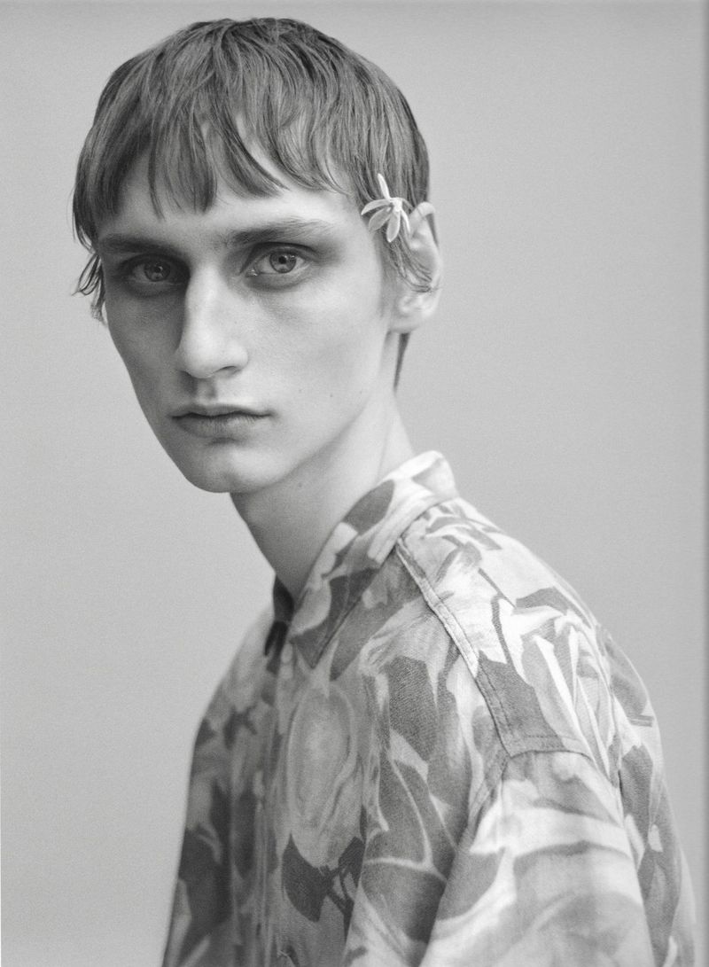 Close-up portrait of a male model wearing a patterned floral shirt with a small flower tucked behind his ear against a plain backdrop.