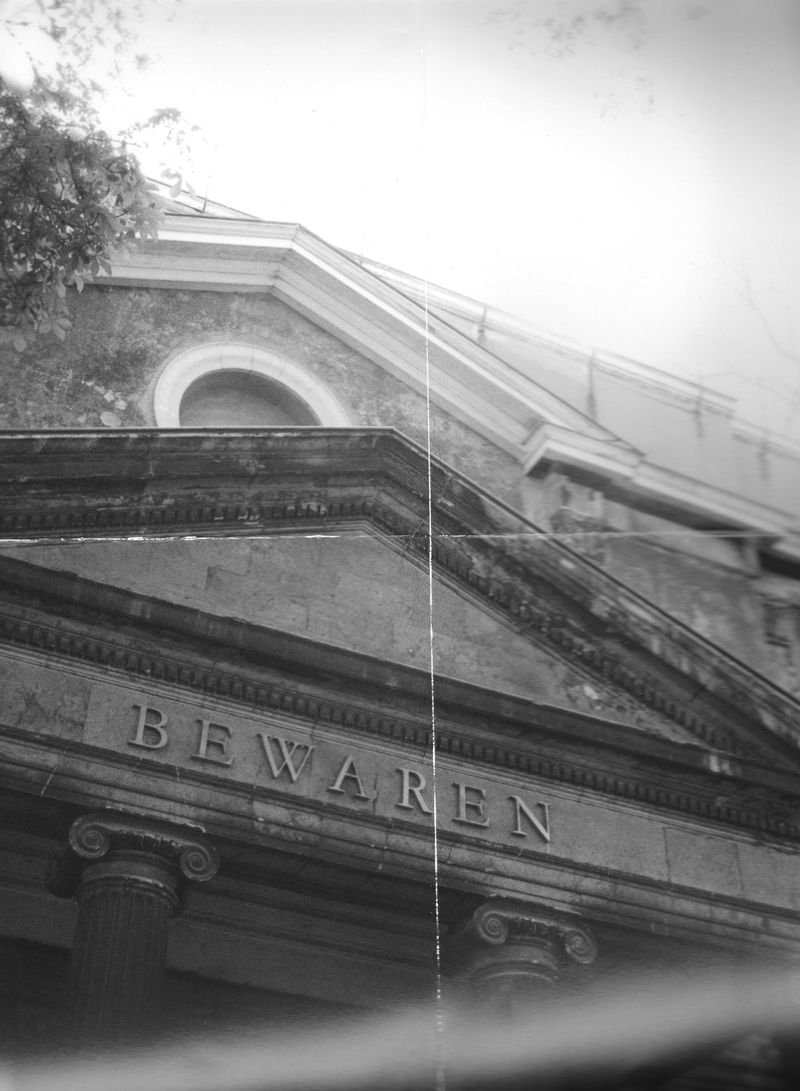 Low-angle monochrome photograph of a classical stone building facade and pediment with the inscription BEWAREN.