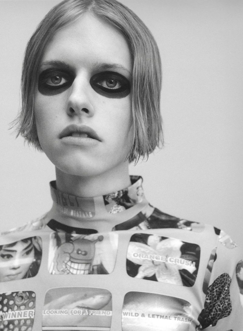 Close-up portrait of a model with heavy dark eye makeup wearing a graphic turtleneck top against a plain studio background.