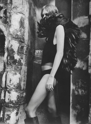 Monochrome fashion portrait of a woman wearing a voluminous black feathered collar and dark bodysuit against a distressed stone wall.