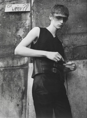 Black and white fashion portrait of a male model in a black sleeveless top and buckled corset belt standing against an aged stone wall.