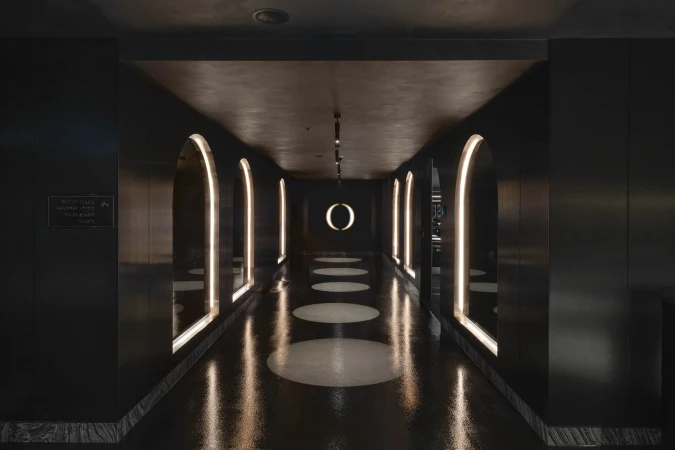Hallway with a symmetrical design featuring dark walls, a reflective floor, and arched, illuminated doorways. Modern interior design.