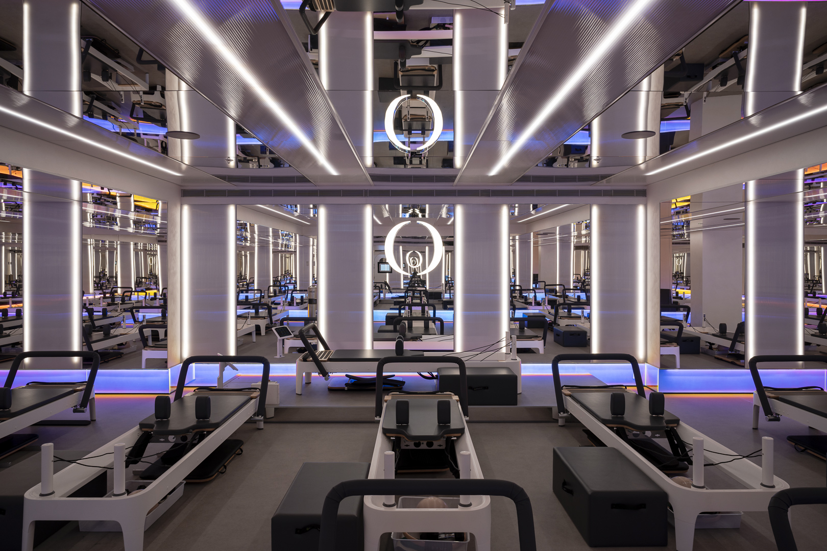 Interior view of a brightly lit, modern fitness studio featuring rowers and exercise equipment. Architectural columns and mirrored ceilings.