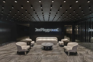 Modern lounge interior featuring white curved armchairs, a matching sofa, and illuminated One Playground signage on a black wall.