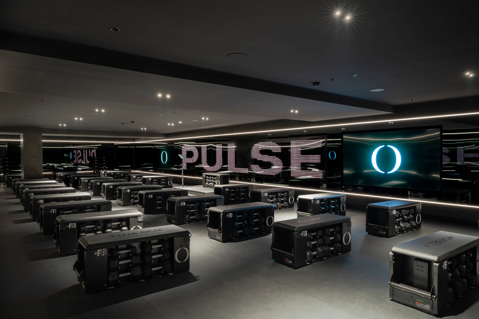 Modern dark fitness studio interior with rows of modular workout benches facing digital screens displaying the word PULSE.