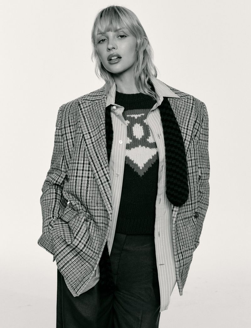 Angèle wearing an oversized plaid blazer, striped collared shirt, and knit sweater against a white seamless studio background.