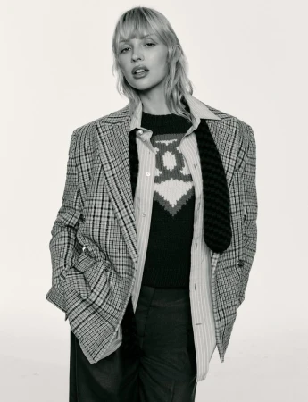 Angèle wearing an oversized plaid blazer, striped collared shirt, and knit sweater against a white seamless studio background.