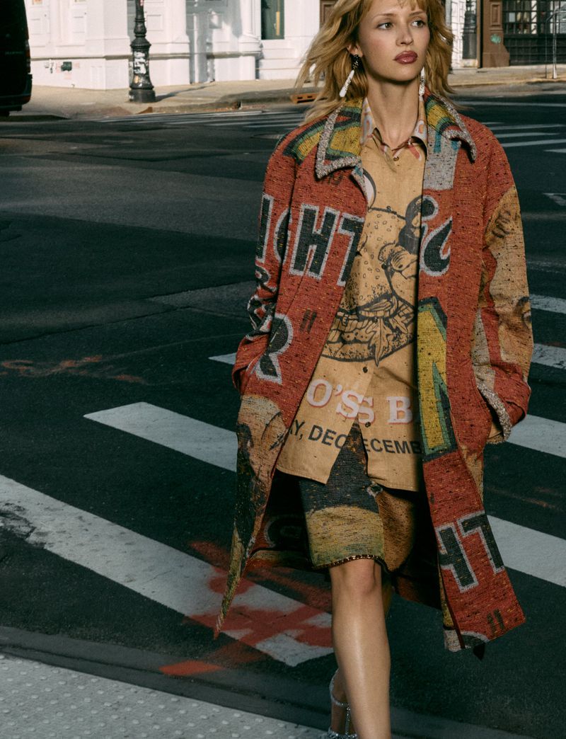Belgian singer Angèle walks across a city street crosswalk wearing a graphic rust and tan printed coat and collared shirt.