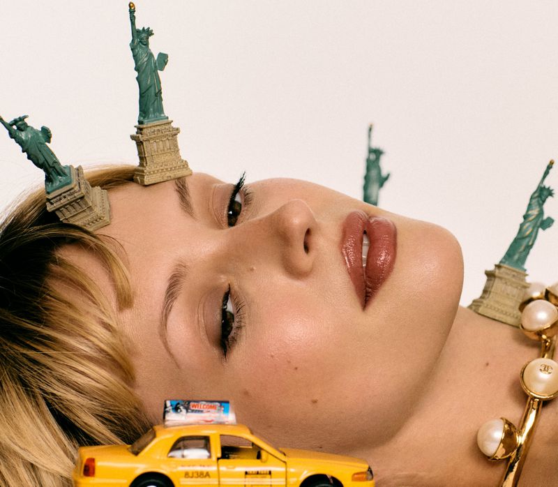 Close-up of a woman lying down with miniature Statue of Liberty figures and a toy yellow taxi on her head and shoulders